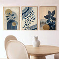 Three framed navy botanical prints with geometric gold and blue accents above a light wood dining table