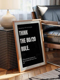 Framed black Pareto Principle poster leaning on floor beside lamp and black leather chair