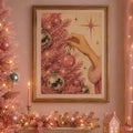Gold-framed pink Christmas tree print with a hand hanging a bauble above a festive mantel