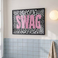 Framed pink SWAG leopard print poster displayed above blue bathroom tile