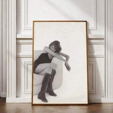 Black-and-white fashion portrait of a woman in knee-high boots seated in an armchair, framed against a white paneled wall