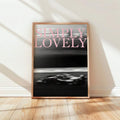 Framed black-and-white racing car poster with pale pink Simply Lovely lettering against a light wall