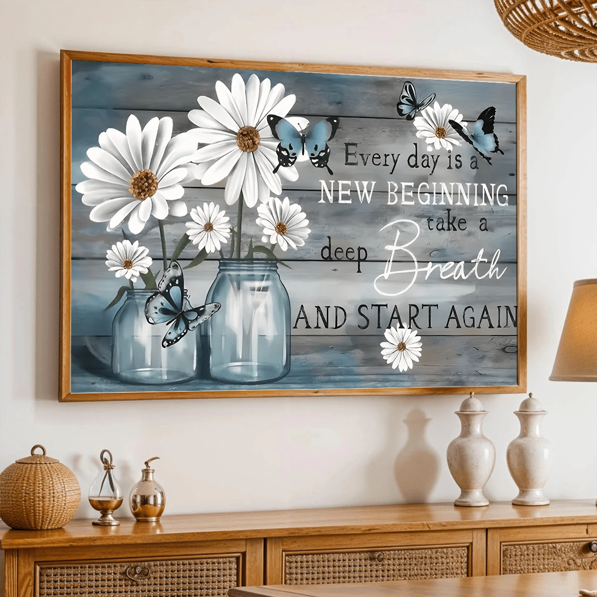 White daisies in blue glass jars with blue butterflies and an encouraging quote in a wood-toned frame above a rattan-front sideboard